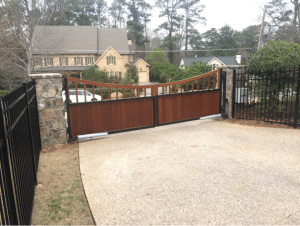 Custom ipe 1x6 driveway gate with arched top design, set between stone columns and black metal fencing in a residential neighborhood.