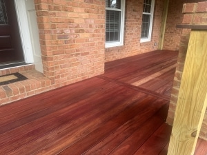 Purpleheart 1x6 porch decking after oil application, highlighting rich deep red and brown tones against a brick home exterior with wood and black metal railing.