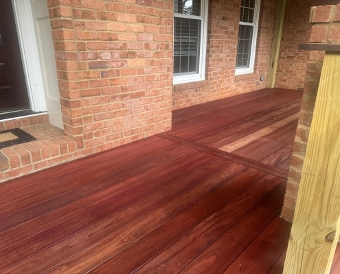 Purpleheart 1x6 porch decking after oil application, highlighting rich deep red and brown tones against a brick home exterior with wood and black metal railing.